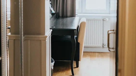 Et træmøbleret arbejdsværelse med skrivebord og stol nær et vindue med gardiner, beige vægge og trægulv.