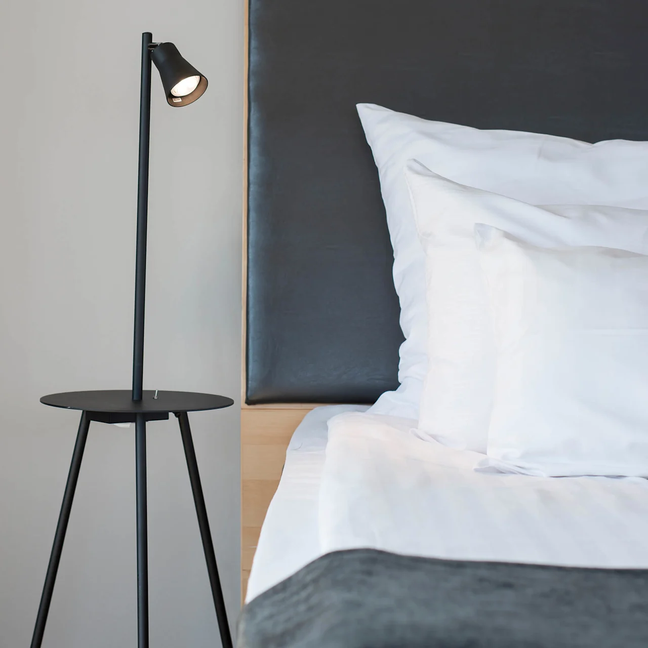 A black floor lamp stands on a modern bedside table, illuminating a neatly made bed with white pillows and sheets, set against a dark headboard.