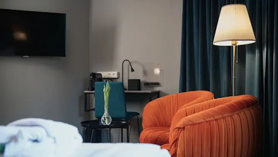 Orange armchairs sit beside a tall floor lamp, facing a small desk with a vase of flowers, against dark curtains and a wall-mounted TV. No text present.