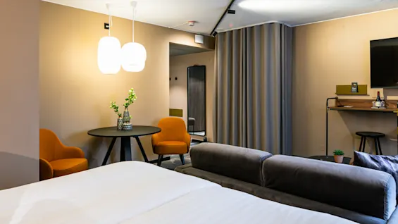 Et moderne hotelværelse med en blidt oplyst dobbeltseng, to orange stole ved et rundt bord med vaser og et vægmonteret TV over et skrivebord med vin og glas.