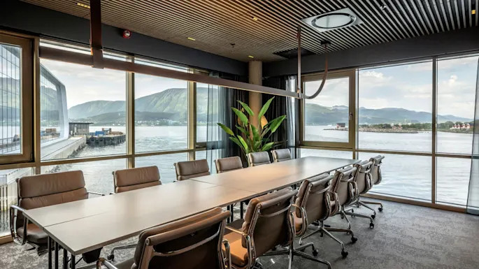 Conference table surrounded by grey chairs, set against large windows overlooking a scenic waterfront and mountains. Indoor plant by the window, wooden ceiling with modern lighting.