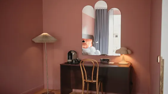 En trästol står vid ett mörkt skrivbord med en kaffemaskin och lampa, mot en rosa vägg. Ovanför speglar som reflekterar en säng med orange sänggavel och gardiner i ett mjukt belyst rum.