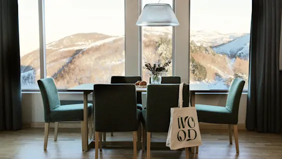 En spisestue med bord og stole, der tilbyder en fantastisk udsigt over sneklædte bjerge. En mulepose med 'WOOD' hænger på en stol.