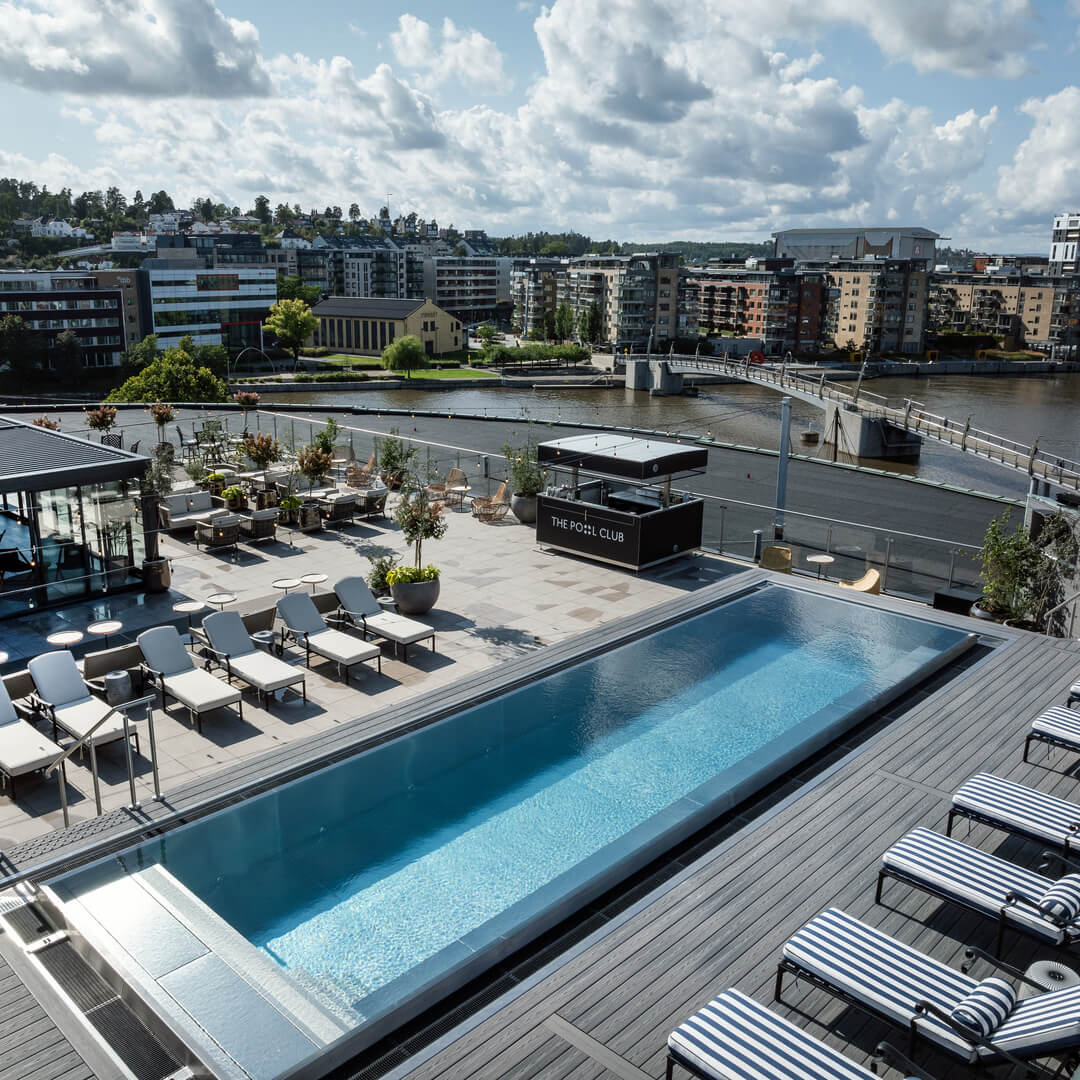 Rooftop swimming pool with surrounding lounge chairs; urban waterfront landscape with buildings in the background.