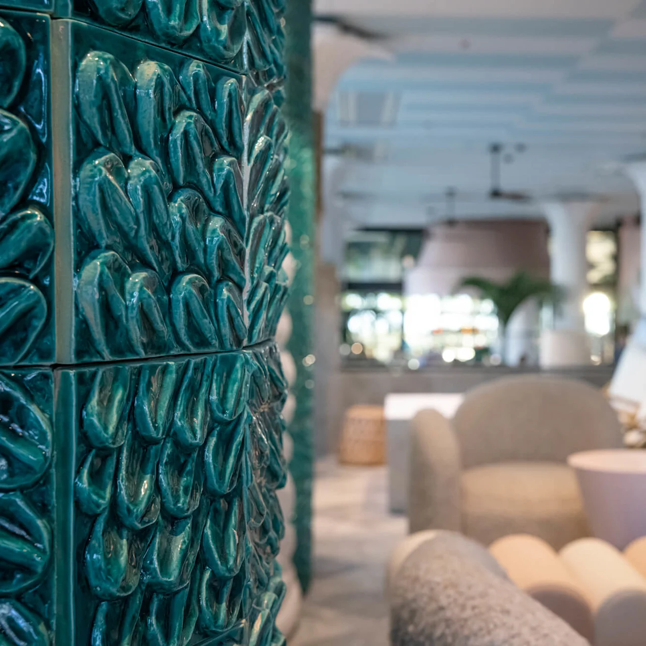 Green textured tile wall; stands prominently. Modern, plush chairs and tables populate the spacious, airy lounge area, with large white columns and soft, inviting lighting in the background.