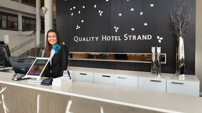 Receptionist smiles behind a desk in a hotel lobby with black wall and white leaf designs. Text reads: "QUALITY HOTEL STRAND" and "LIFE IS AN ADVENTURE."
