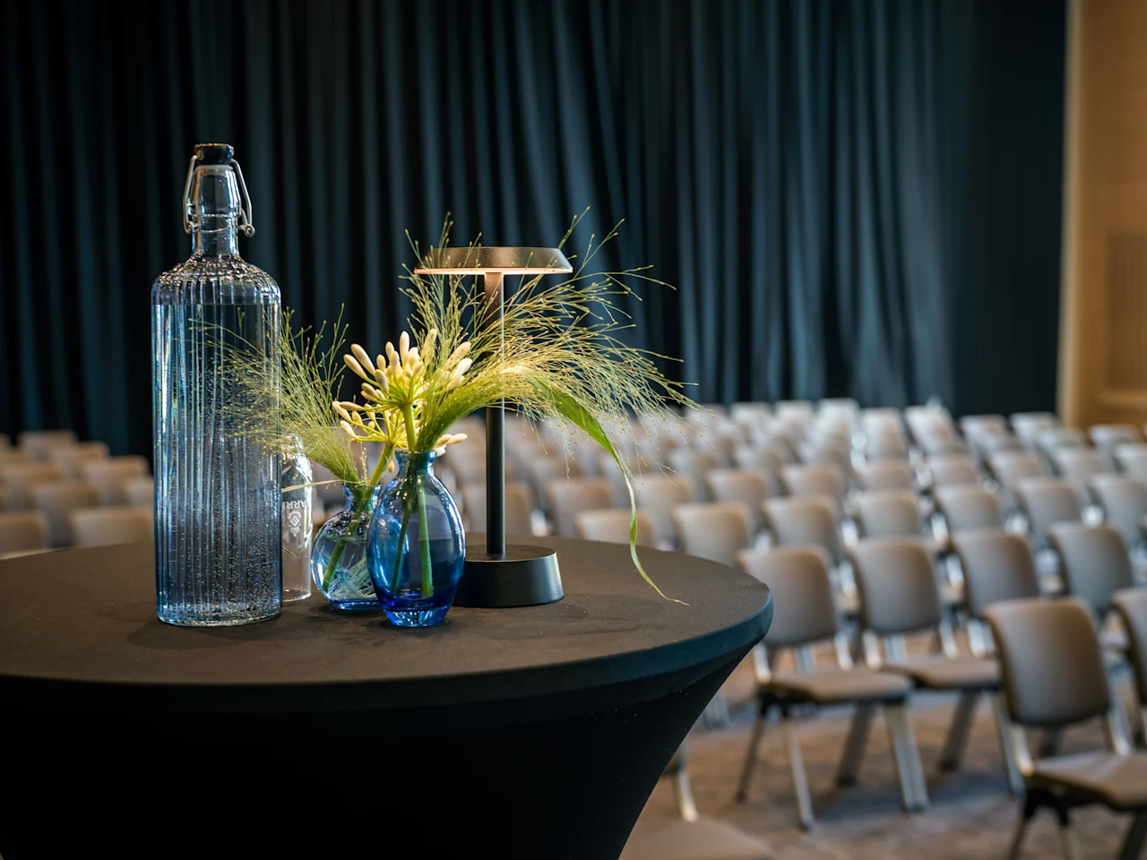 Ett bord med en vattenflaska och blomsterdekoration i ett konferensrum med rader av tomma stolar och en mörk gardin.