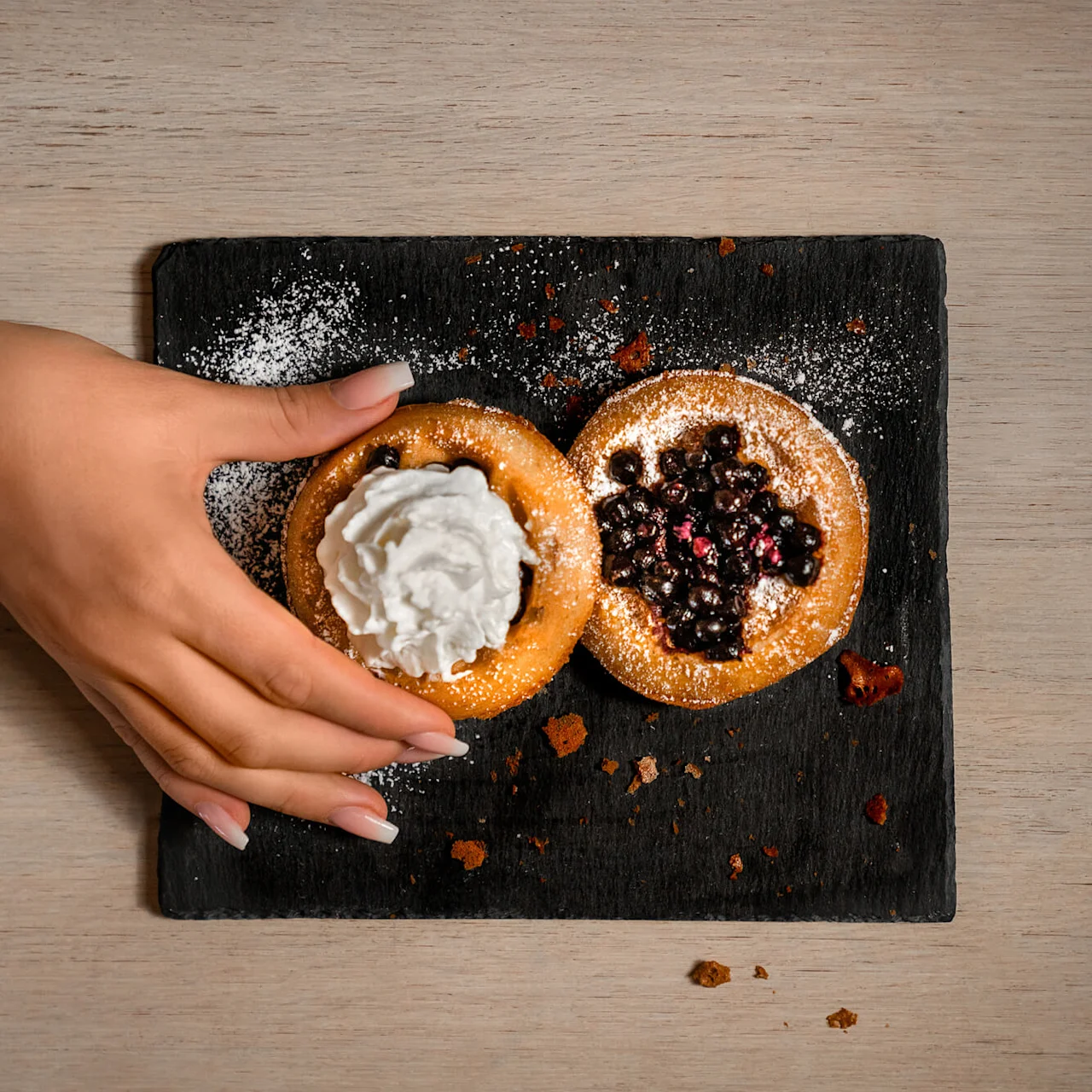 A hand is taking a pastry with whipped cream from a slate board with two pastries, on a wooden table.