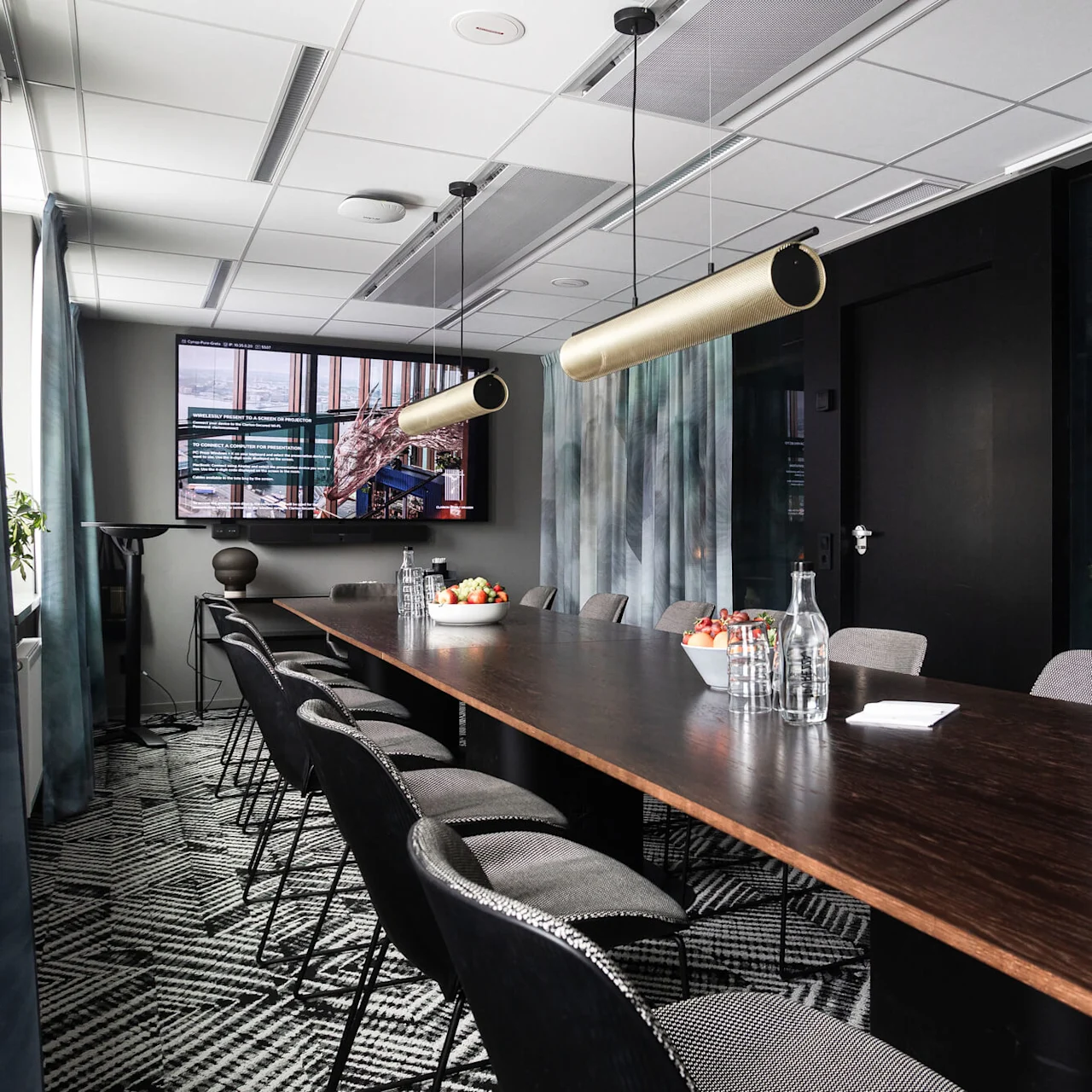 Konferensbord med stolar och fruktskålar, placerade under moderna taklampor. Väggarna är smyckade med gardiner och en stor skärm som visar texten "HYLLIE TERRASS."