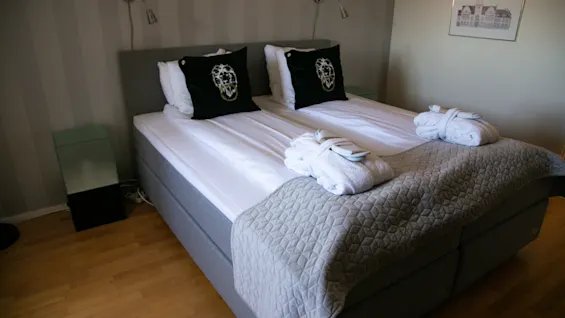 A cozy double bed in a hotel room with white bathrobes and dark decorative pillows.