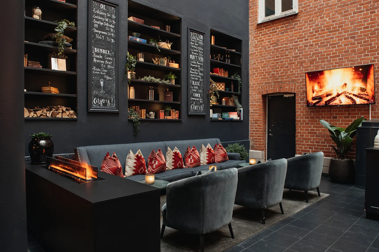 En hyggelig lounge med en moderne pejs, komfortable siddepladser og bogreoler. Et TV viser en pejs, med tavler der siger enjoy og cheers.