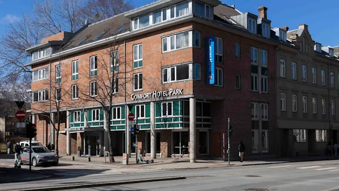 Kolmikerroksinen tiilirakennus hotelli suurine ikkunoineen sijaitsee kaupunkikadulla, lähellä on useita lehtipuita. Kyltissä lukee “COMFORT HOTEL PARK” ja “COMFORT HOTEL” pystysuorassa kyltissä.