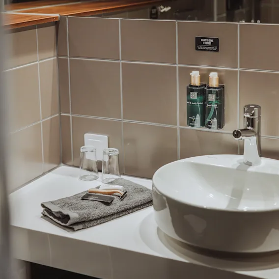 En hvit, rund servant med en elegant kran står på en benk. Nær ligger foldede håndklær, glass og monterte toalettsaker mot beige flislagte vegger. Tekst: "SOOQ VENTURELLA".
