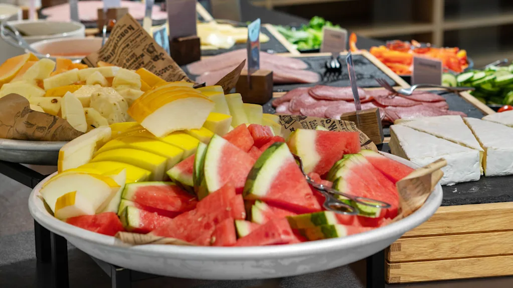 Slices of various fruits such as watermelon, melons, and pineapples are arranged on a serving platter alongside deli meats and cheese on a buffet table.