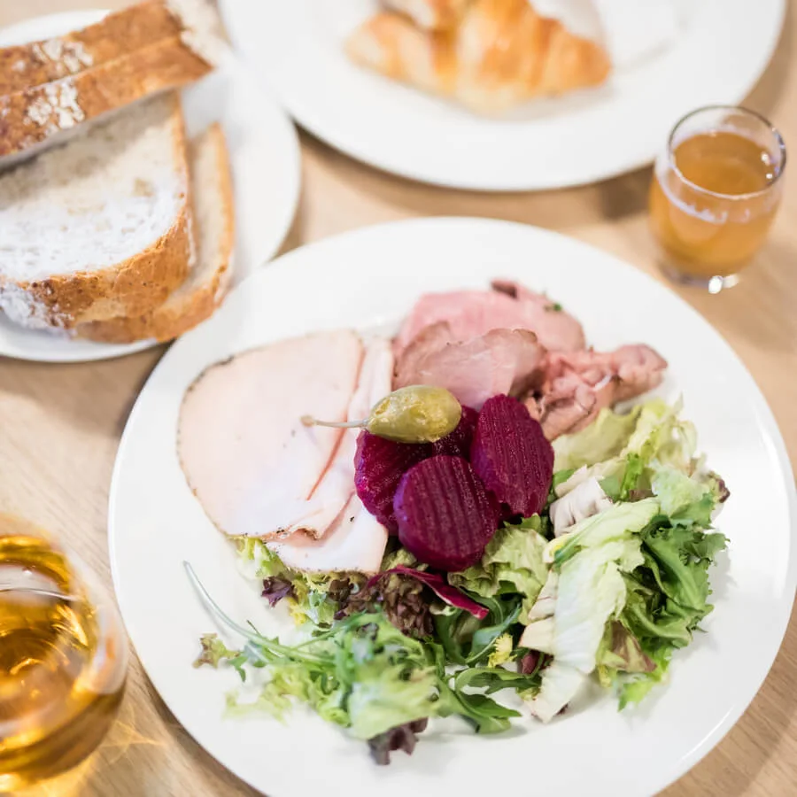 Ett fat håller skivad kött, rödbetor och sallad, nära brödskivor, en croissant med druvor och drycker. Allt är arrangerat på ett ljust träbord.