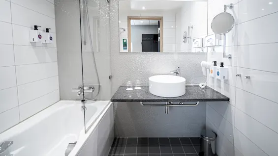 A bathroom features a white circular sink on a black countertop with a mirror above it. A bathtub with a glass screen is next to it, and toiletries are wall-mounted.