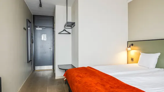 Et koselig hotellrom med en ryddig seng med et oransje teppe. En myk lampe lyser ved siden av, og skaper en varm atmosfære ved den enkle sorte døren og minimalistiske møbler.