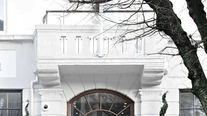 A wooden double door, adorned with a Christmas wreath, stands between two spiral topiary plants in black pots labeled "OLEANA". The white building has large windows and a balcony.