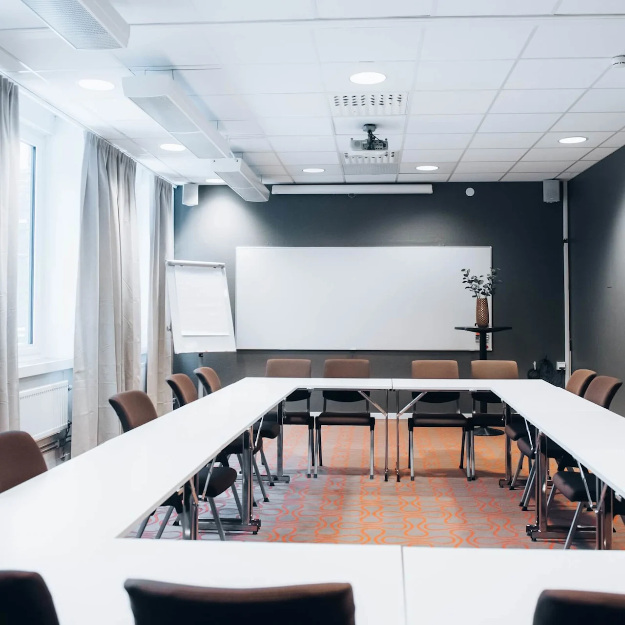 Meeting room features a U-shaped table arrangement surrounded by chairs. In the background, a whiteboard and flip chart stand before a window with beige curtains. A small potted plant decorates the room.