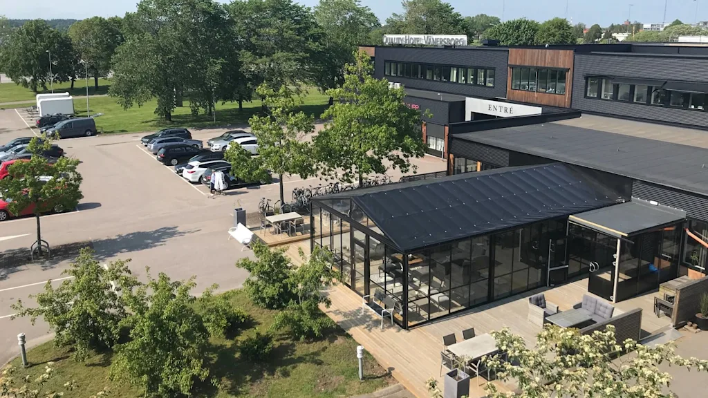En modern byggnad märkt "Quality Hotel Vänersborg" med en glasutbyggnad står bredvid en parkeringsplats full av bilar. Omgivande träd och gräsmattor förbättrar den urbana miljön.