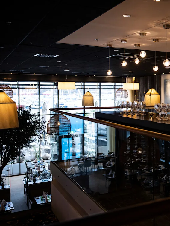 Glasses arranged on a bar counter under modern pendant lights. The setting is a dimly lit, upscale restaurant with large windows and chic decor.