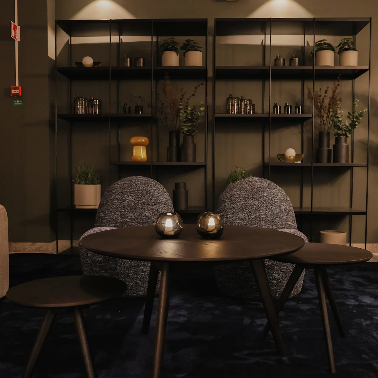 Two dark round tables sit in front of plush chairs, surrounded by beige sofas and leafy plants, set against a backdrop of shelves with decorative items in a warmly lit room.
