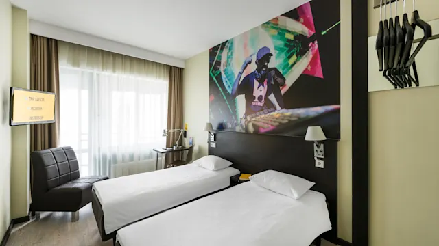 Two neatly made single beds face a wall mural of a DJ in a modern hotel room. A television displays options: "TRIP ADVISOR," "FACEBOOK," and "INSTAGRAM." A chair and desk with lamp are nearby.