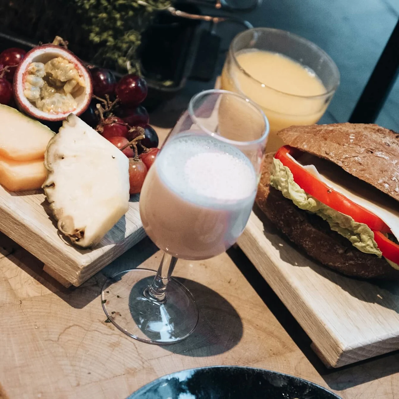En sandwich med salat, tomat og ost på en træplade, omgivet af frugt, et glas milkshake og kopper med juice og kaffe, placeret på et træbord.
