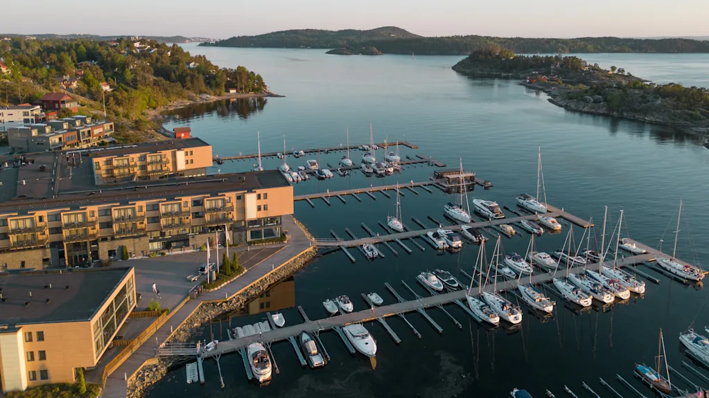 Både fortøjet ved en marina, omgivet af roligt vand. Bygninger omkranser vandkanten, mod en baggrund af frodig grønt og små øer under en klar himmel.