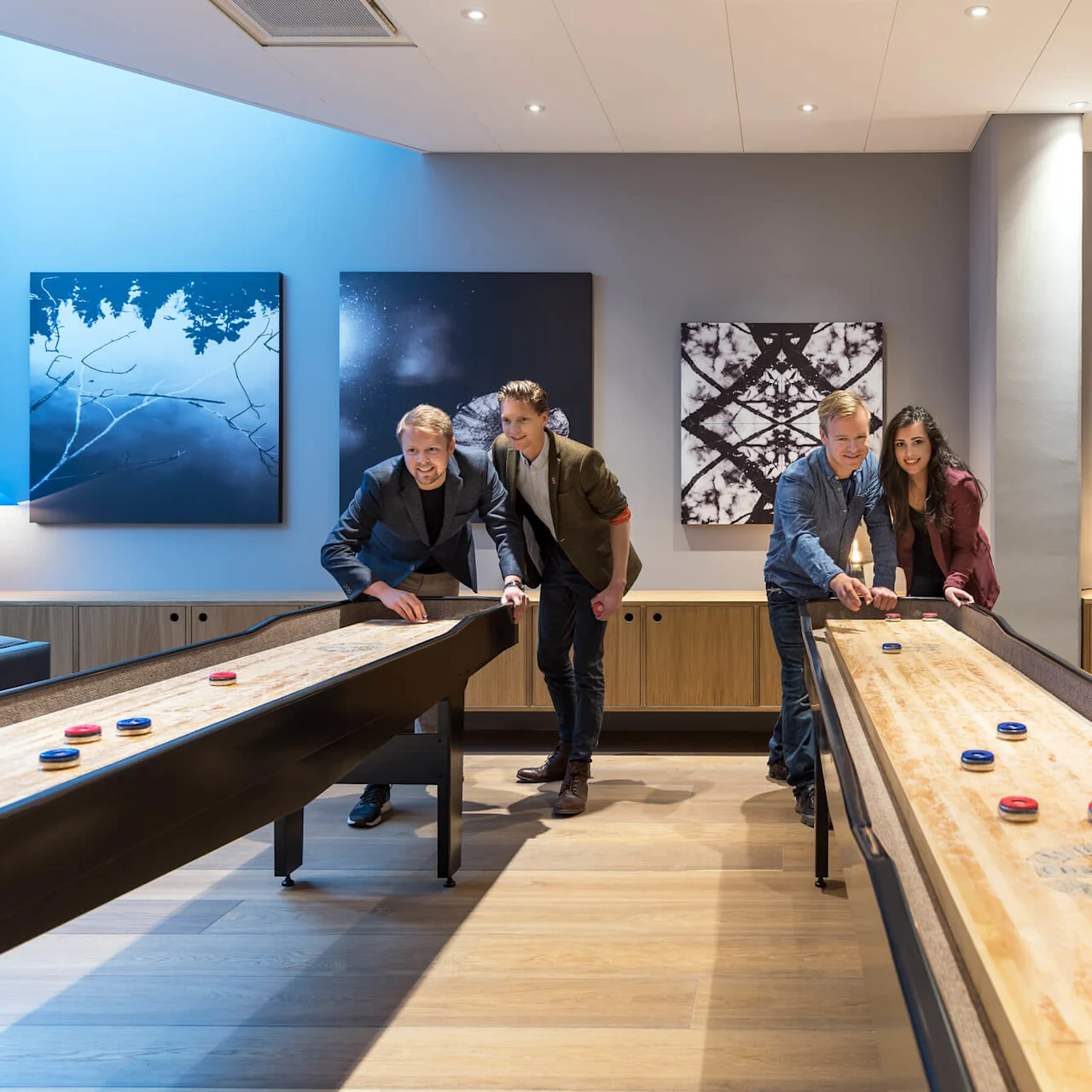 Four people play shuffleboard in a modern room. The wooden floor and stylish furniture, including paintings and lamps, create a cozy atmosphere.