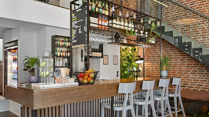Bar counter with high chairs, displaying bottles and glasses on shelves. Menu board lists: Snacks, Bites, Beer & Cider, Wine, Beverages, Snacks, and Sweets. Brick wall and indoor plants surround. 