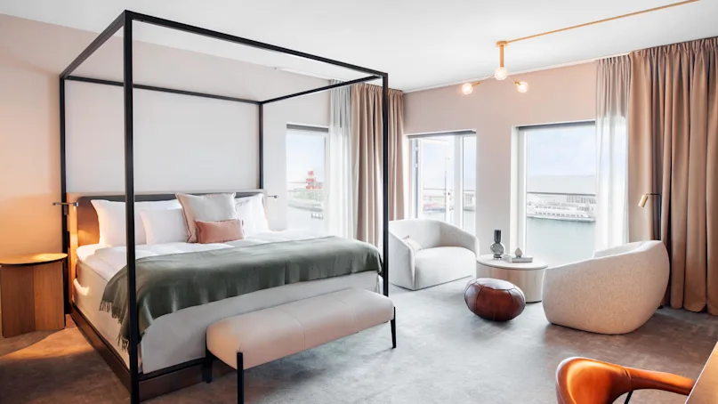 A spacious bedroom features a large canopy bed with green bedding, surrounded by modern chairs and a side table. Large windows offer a view of the sea, enhancing the serene setting.