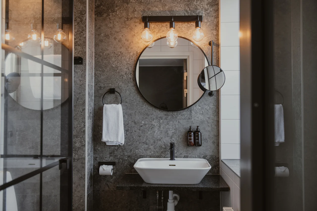 Ett modernt badrum med handfat, rund spegel och belysning i industristil. En vit handduk hänger i närheten.