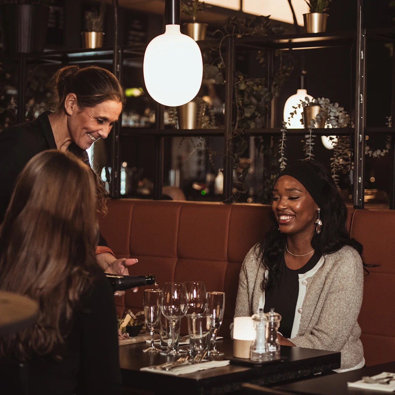 A waiter chats with two women seated at a restaurant table, smiling and surrounded by glasses and napkins, with hanging lamps and decorative plants creating a cozy atmosphere.