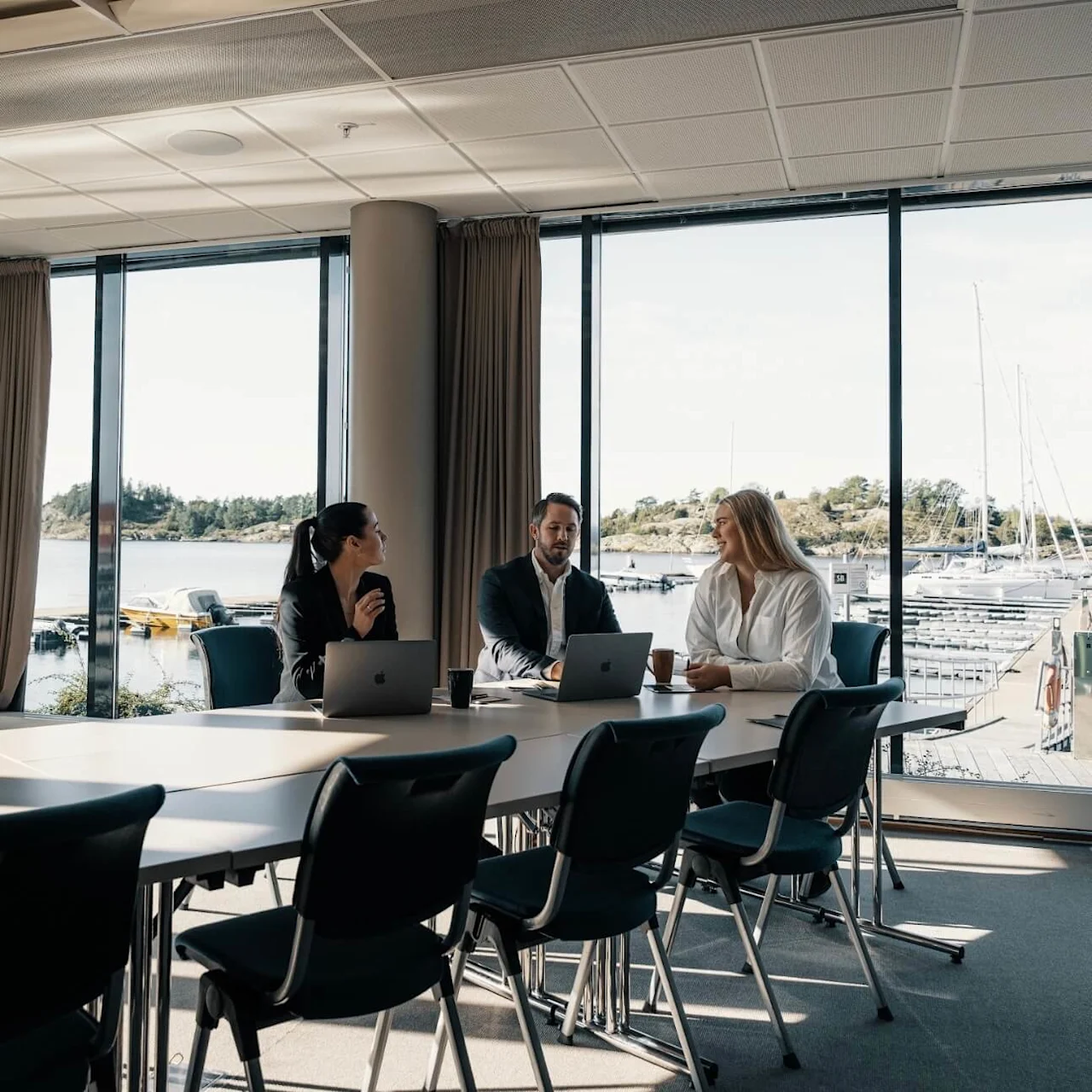 Tre personer sitter rundt et bord med bærbare datamaskiner og snakker sammen. De er i et lyst rom med store vinduer som vender mot en marina full av båter.