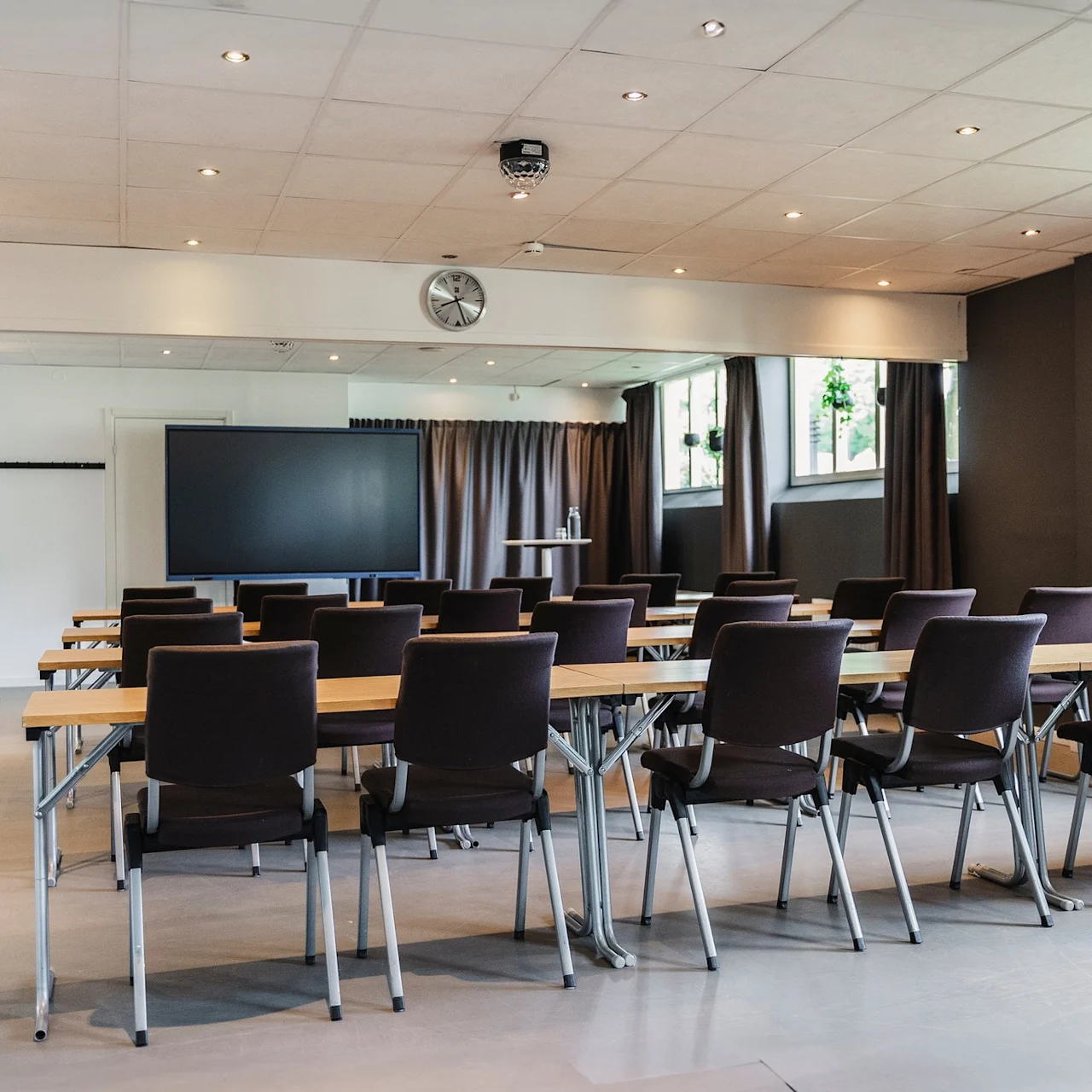Rader av tomma stolar och bord vetter mot en stor svart skärm i ett konferensrum, omgiven av beige väggar och fönster med mörka gardiner.