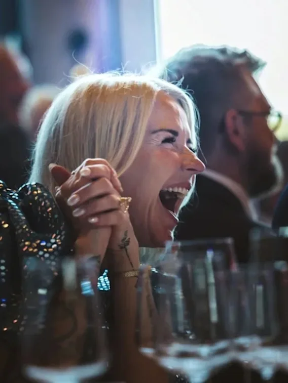 A blonde woman laughing joyfully with clasped hands at a dinner table filled with wine glasses during a part