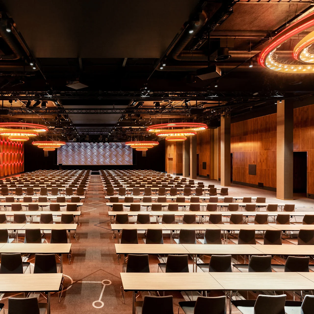 Rows of tables and chairs are arranged in a large, modern conference hall with wooden walls, red lighting accents, and circular chandeliers. A large, empty screen is displayed at the front.