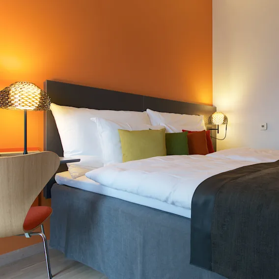 Et koselig hotellrom har en pent oppredd seng med fargerike puter, opplyst av varme nattbordslamper, satt mot en oransje aksentvegg.