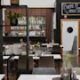 En restauranginteriör har bord med stolar, växter och en bakvägg med hyllor av flaskor. Ovanför en dörr står en skylt med texten "HERE IS WHERE THE MAGIC HAPPENS."