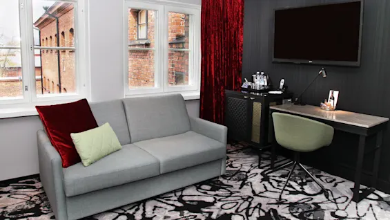 A modern living room with a gray sofa, a work desk, and a wall-mounted TV. Windows show a brick building outside.