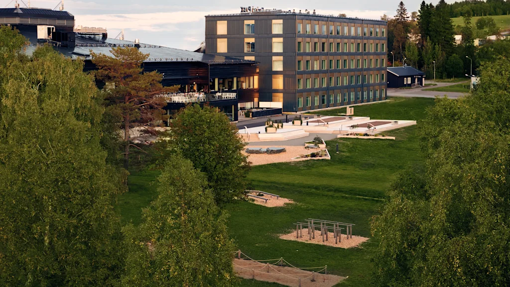 En moderne, fleretasjes bygning står ved siden av en frodig grønn park med forskjellige utendørs treningsapparater, omgitt av store trær. Himmelen er delvis skyet, som antyder en rolig sen ettermiddag.
