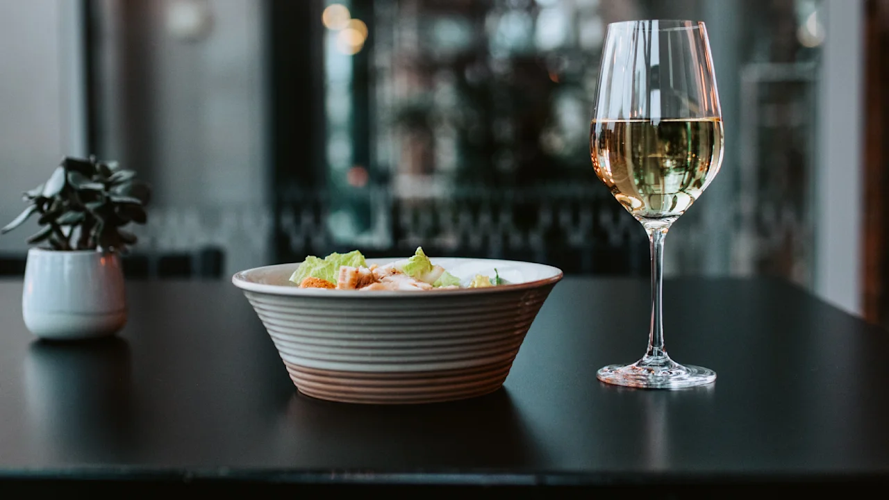 En skål med sallad bredvid ett glas vitt vin på ett svart bord i ett modernt matsalsområde med glasväggar.