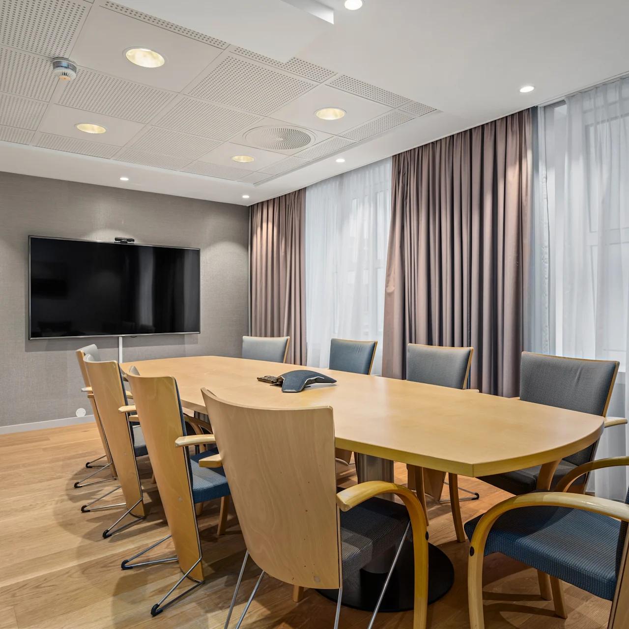 Et stort konferencebord i træ omgivet af stole, vendt mod en vægmonteret skærm i et lyst rum med gardiner. En telefon står på bordet.