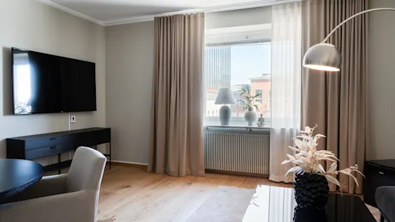 En modern vardagsrum har en väggmonterad TV, ett elegant svart skåp och en bekväm fåtölj. Gardiner ramar in ett fönster med en lampa och växt, beläget i ett ljus rum.