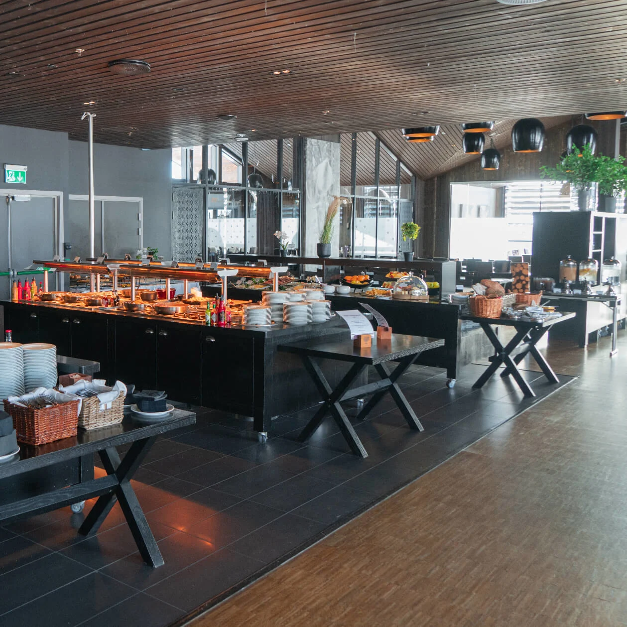 Buffet tables laden with various dishes, plates, and utensils, arranged in a spacious dining area. A wolf photograph and green plants decorate the walls, creating a modern, elegant ambiance.