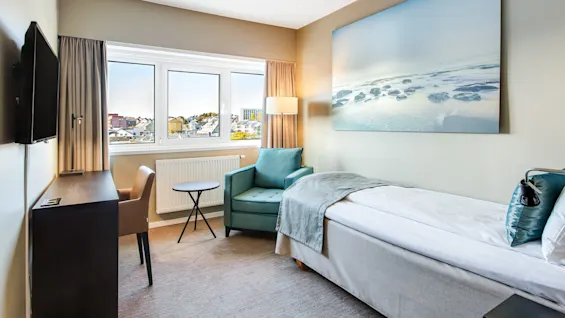 Et ryddig hotellrom viser en enkeltseng med turkise detaljer. Et maleri av en kystscene pryder veggen. Et vindu viser en urban byutsikt under en klar himmel.