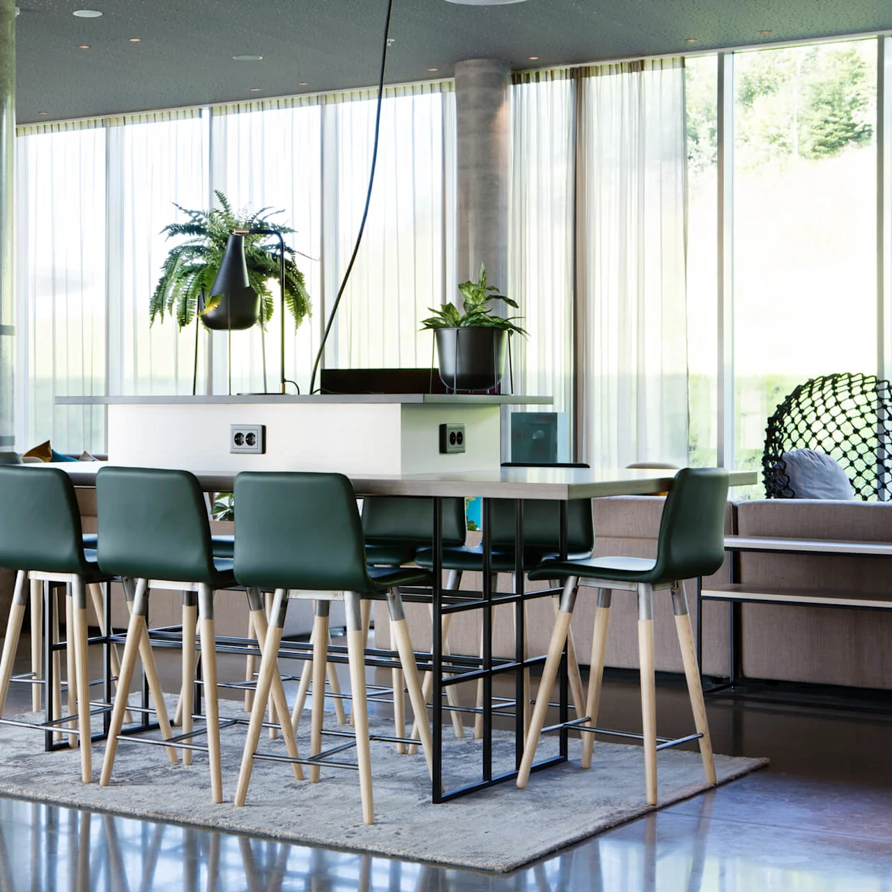 Dining table with green chairs and plants, situated in a modern living area with art on the wall. Large windows allow natural light, creating an airy atmosphere.