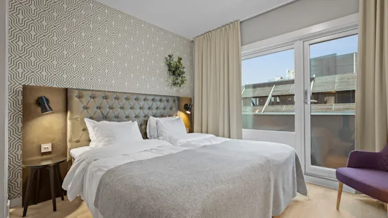 Ett mysigt hotellrum med en stor säng, mönstrad tapet och ett fönster med utsikt över närliggande byggnader. En lila stol står vid fönstret.