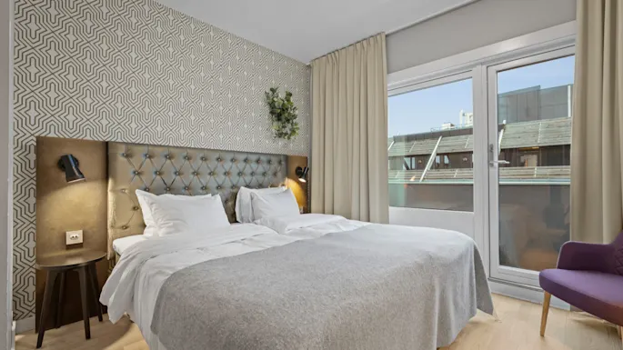 Et koselig hotellrom med en stor seng, mønstret tapet og et vindu som tilbyr utsikt over nærliggende bygninger. En lilla stol står ved vinduet.